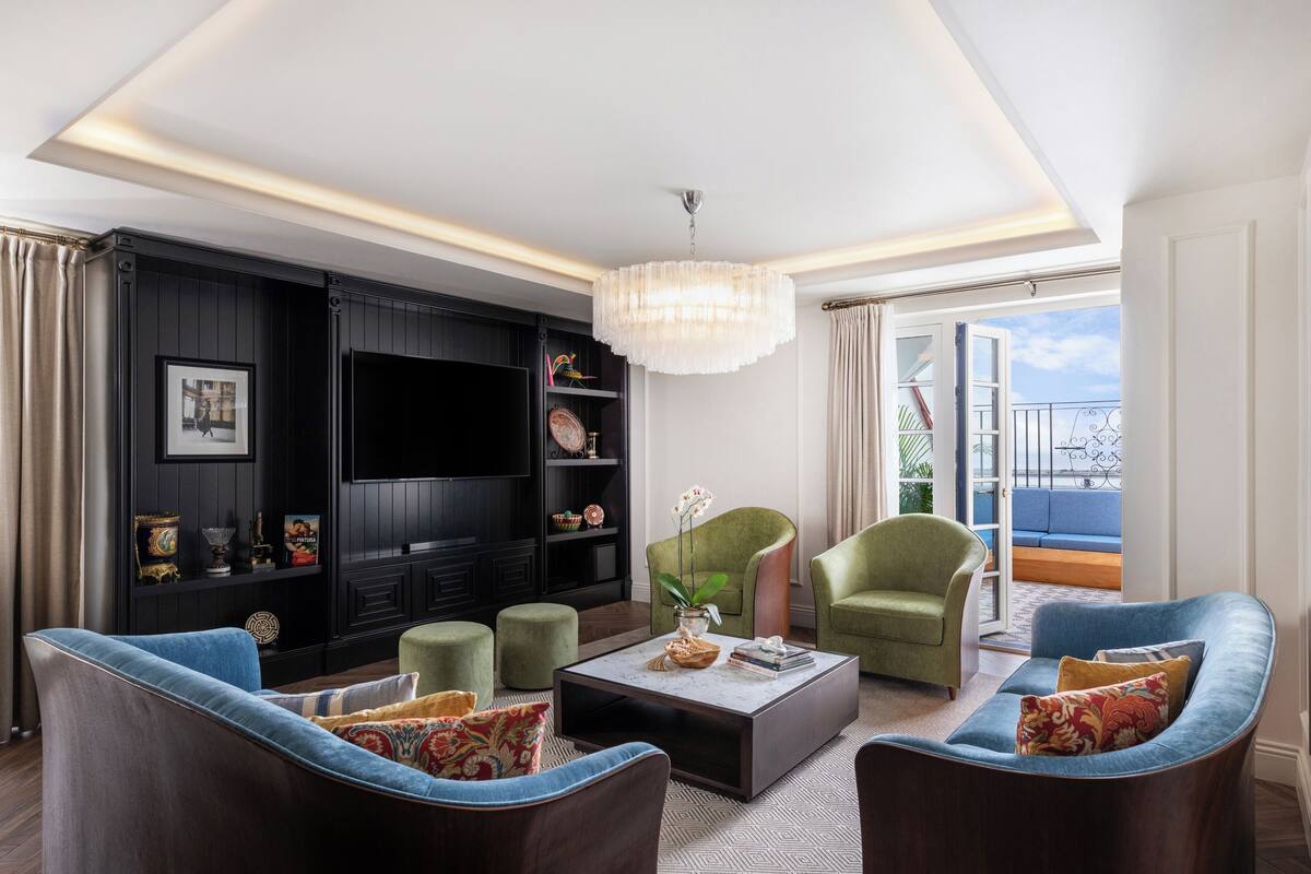 A modern living room with a flat-screen TV, green armchairs, a coffee table, and a balcony with a view.