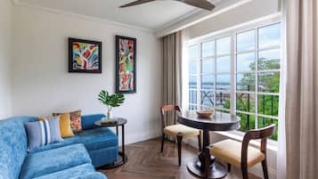 A modern living room with a blue sofa, two framed abstract paintings, a dining table with chairs, and a view of greenery through large windows.