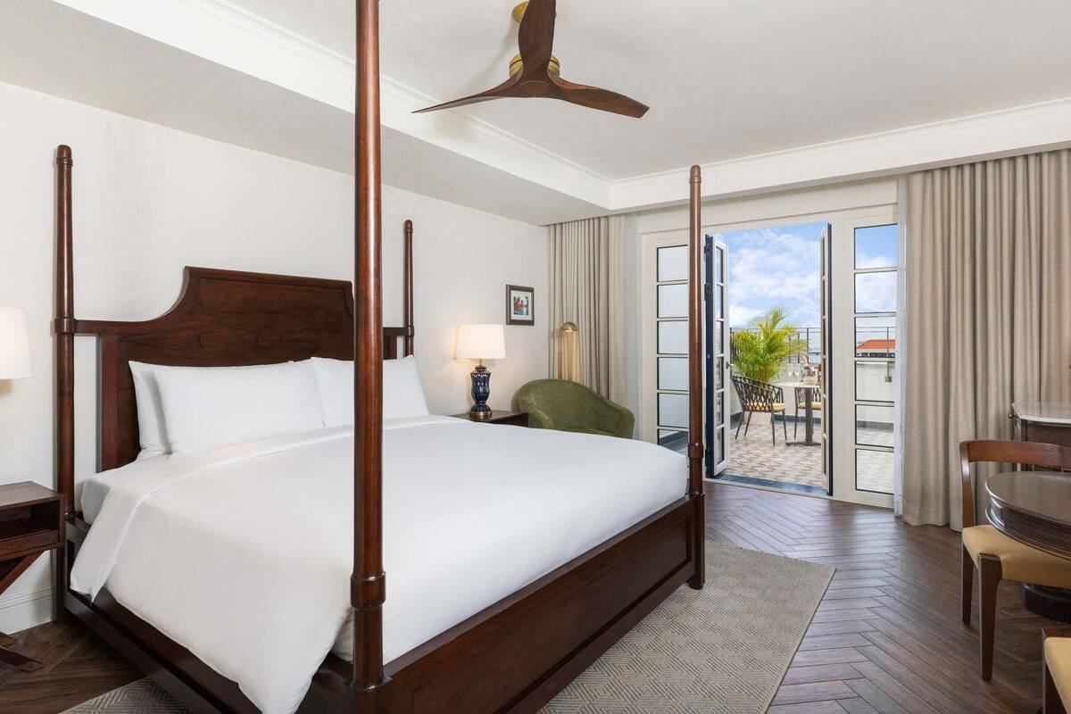 A hotel room with a four-poster bed, a ceiling fan, a balcony with a view, and a dining area with a table and chairs.