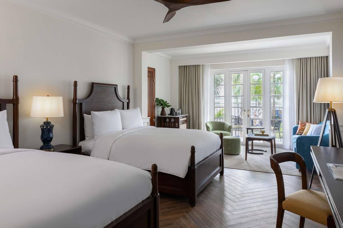 A hotel room with two beds, a ceiling fan, a balcony with a view, and a dining area with a table and chairs.