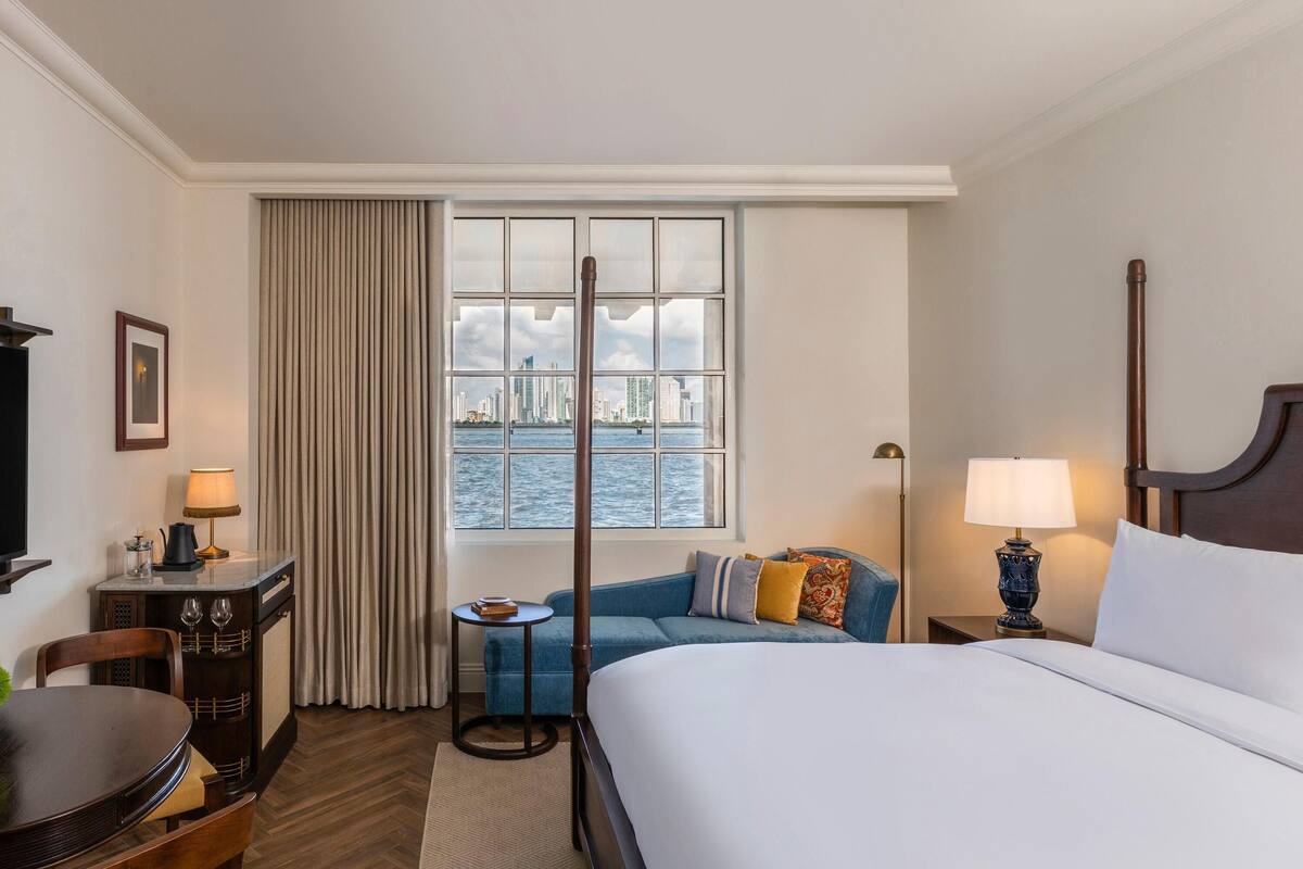 A hotel room with a large bed, a desk with a lamp, a chair, a small table with a lamp, a sofa with cushions, a window with a view of a cityscape, and a curtain.