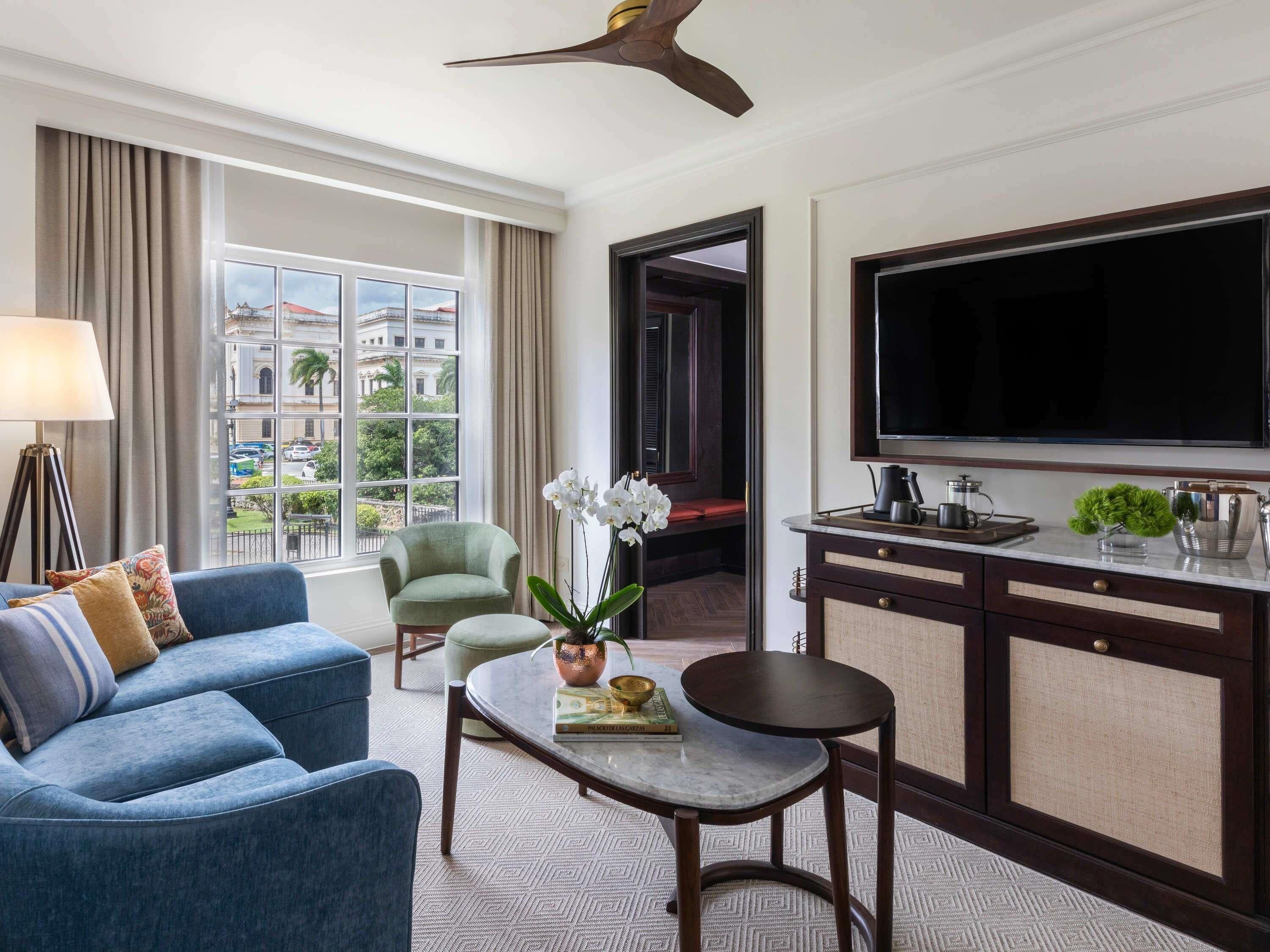 Ein Hotelzimmer mit einer Couch, einem Sessel, einem Couchtisch, einem Fernseher und Blick auf Gebäude durch das Fenster.