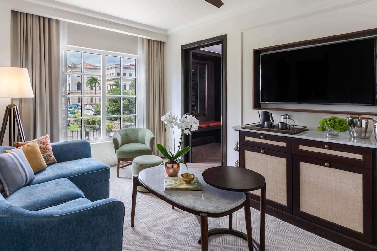 Ein Hotelzimmer mit einer Couch, einem Sessel, einem Couchtisch, einem Fernseher und Blick auf Gebäude durch das Fenster.