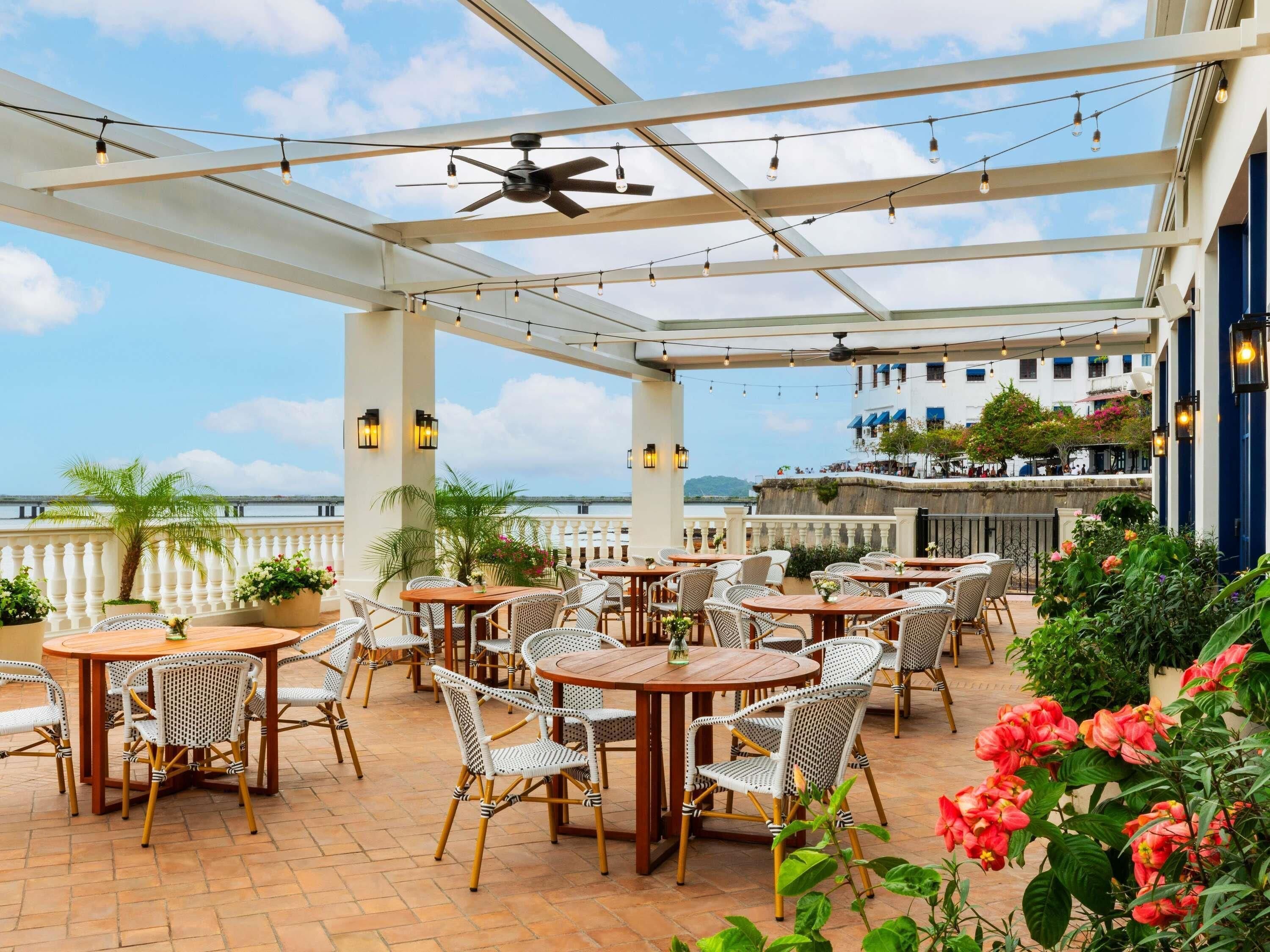 An outdoor patio with round tables and wicker chairs, overlooking a body of water.