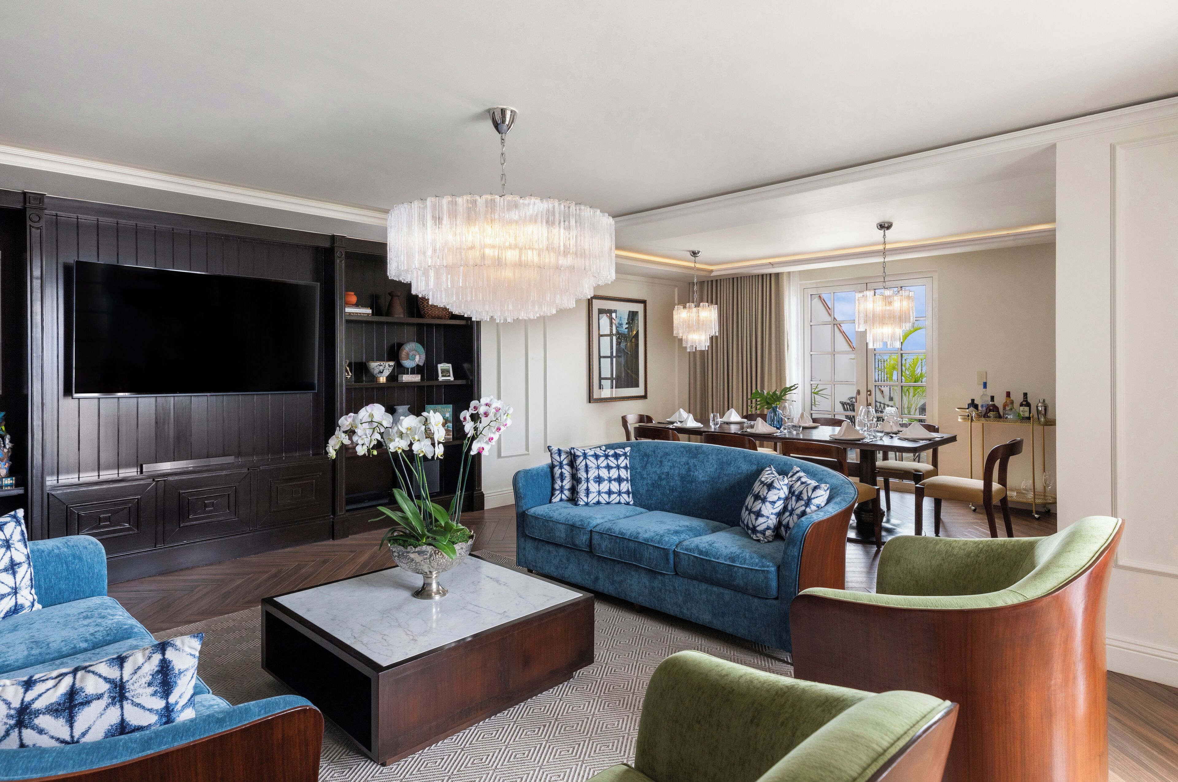 A modern living room with a large flat-screen TV, a blue sofa, a coffee table, and a chandelier.