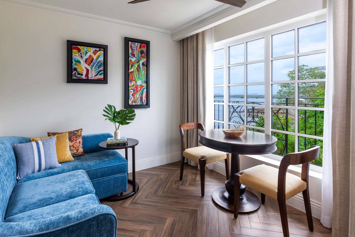 A modern living room with a blue sofa, two framed abstract paintings, a round table with chairs, and a view of a body of water through large windows.