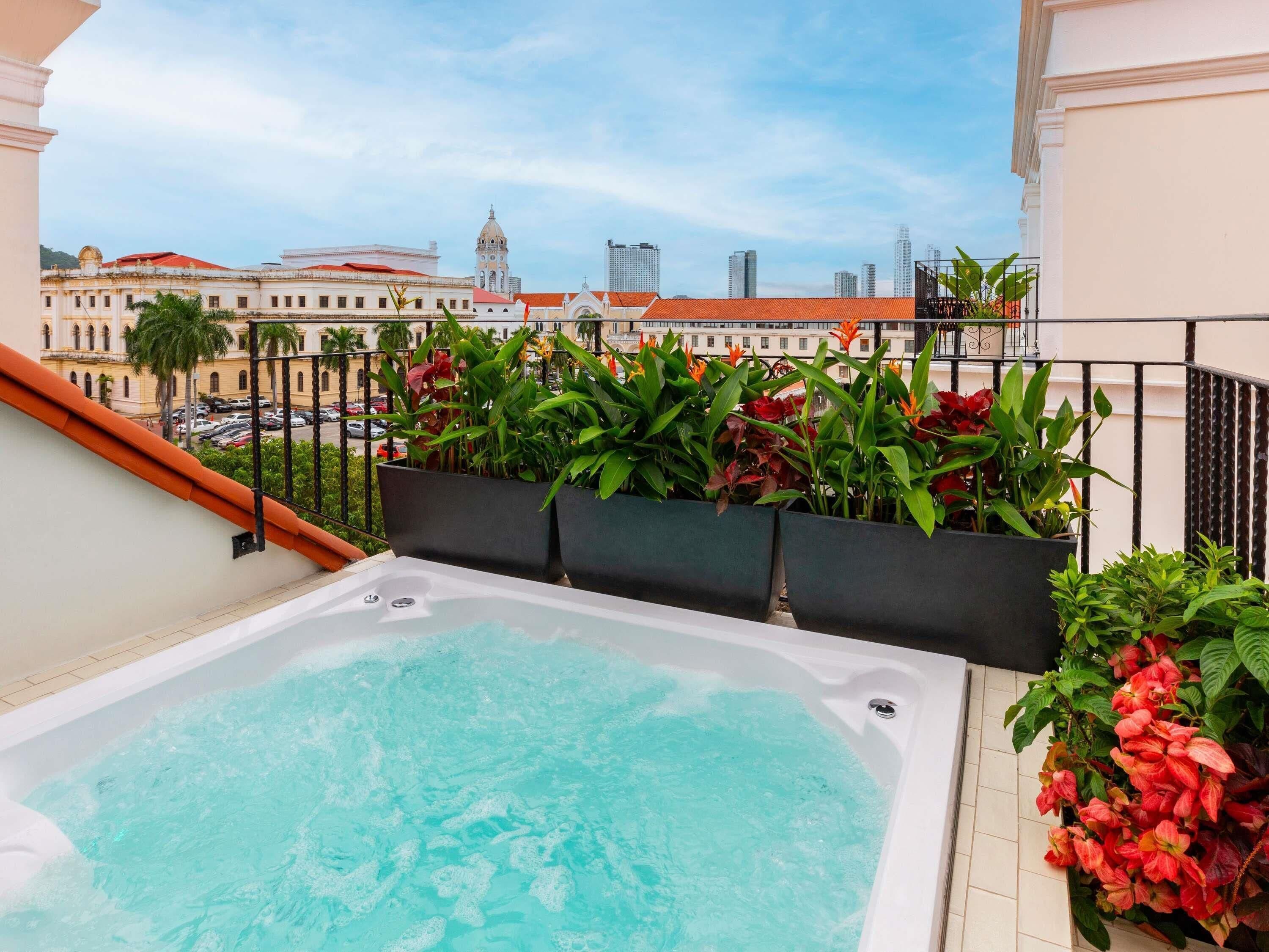 Un jacuzzi en la azotea con vistas a la ciudad, rodeado de plantas en macetas y una barandilla de balcón.