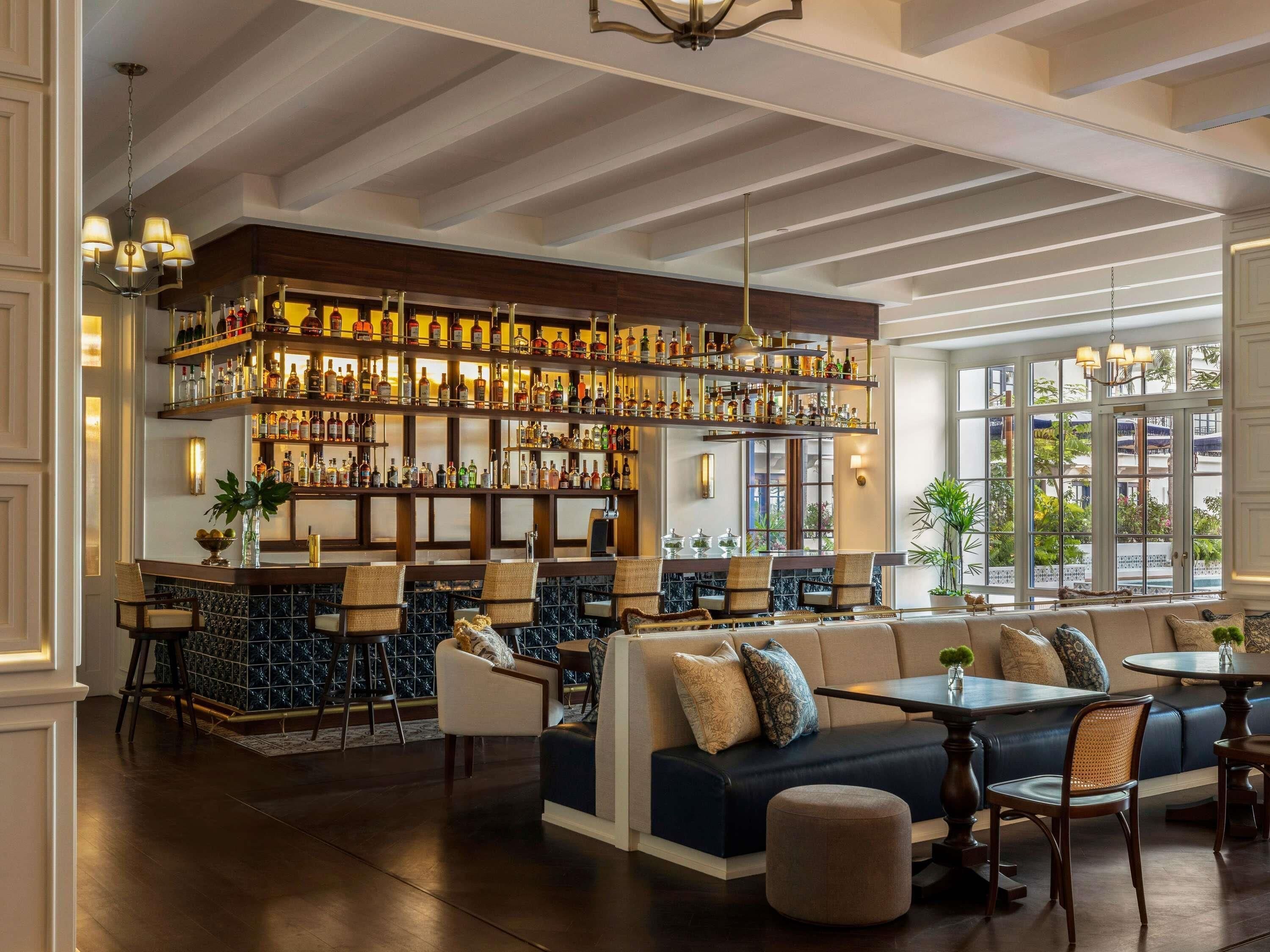 A bar area with a high ceiling, wooden beams, and a variety of liquor bottles. There is seating with a sofa and chairs, and a large window providing a view of the outdoors.