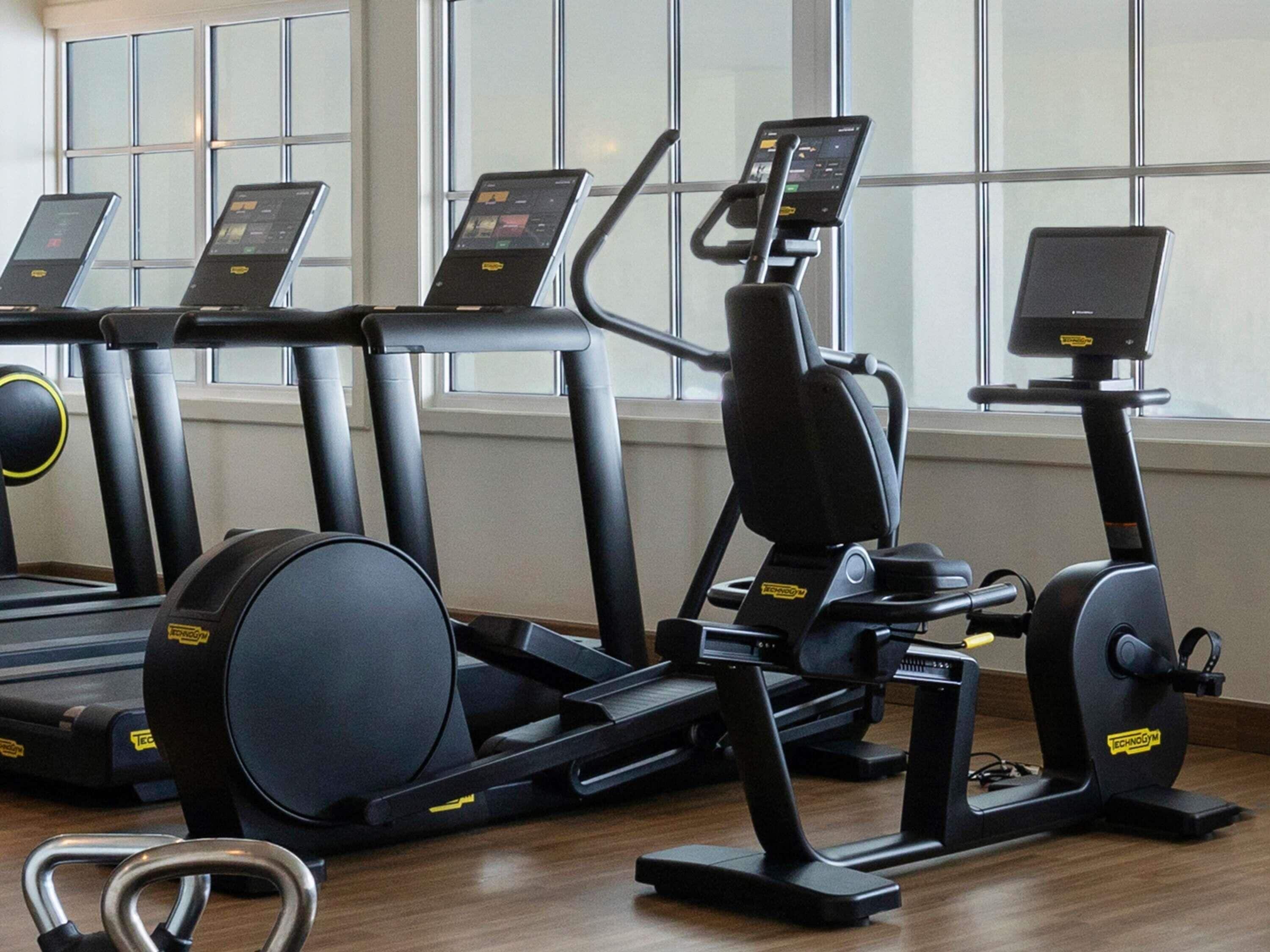 A gym with treadmills, elliptical machines, and a stationary bike, with large windows in the background.