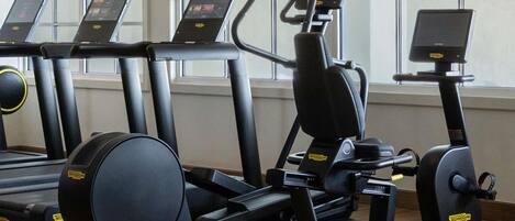 Un gimnasio con cintas de correr, elípticas y una bicicleta estática, con grandes ventanales al fondo.