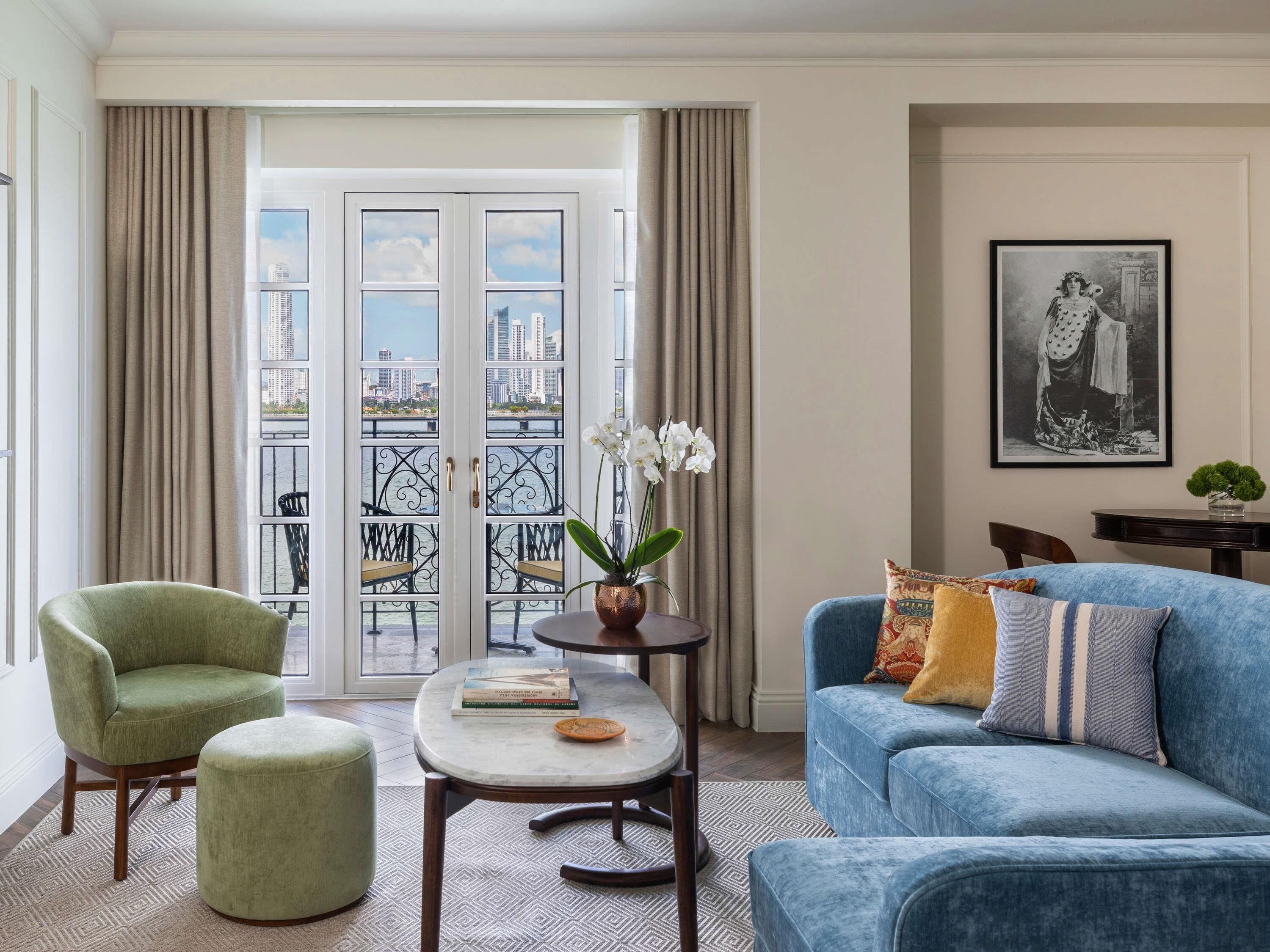 Un salon avec un fauteuil vert, un canapé bleu, une petite table avec des livres et une vue sur le paysage urbain, visible à travers une porte vitrée.