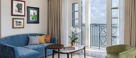 A living room with a blue sofa, a round table, a green armchair, and a balcony with a railing.