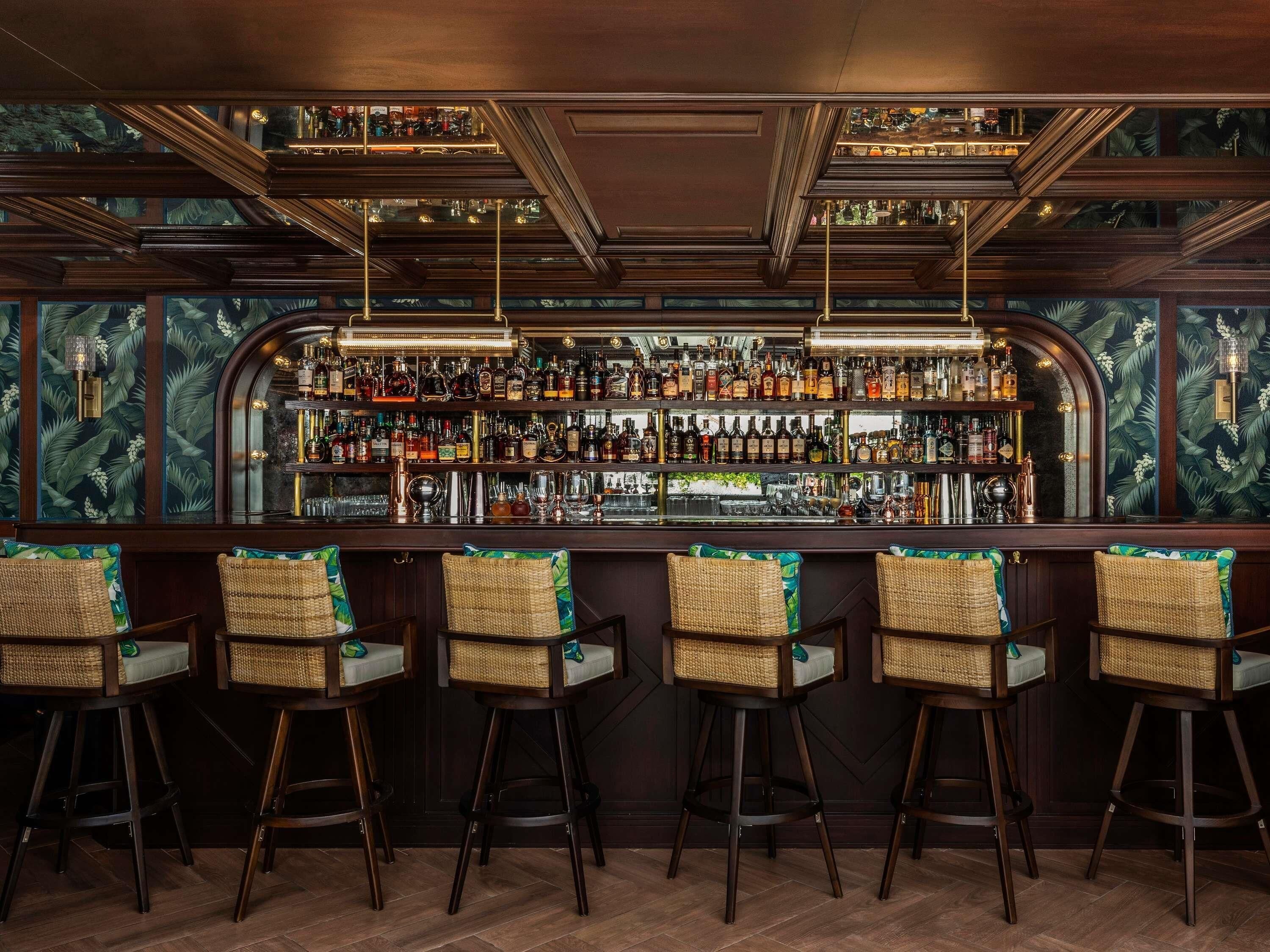 A bar area with a wooden bar counter, multiple shelves of bottles, and wicker bar stools.