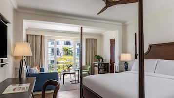 A hotel room with a four-poster bed, a desk with a lamp, a chair, a small table with a plant, and a view of the outside balcony.