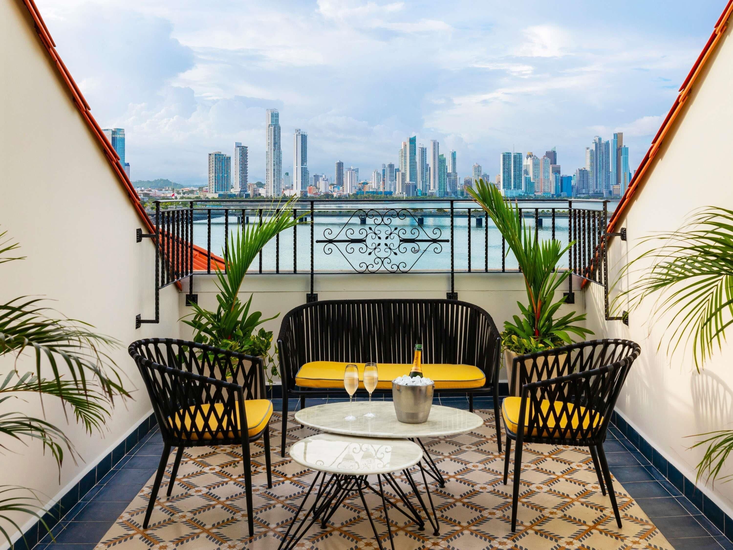 A balcony with a city view, two black chairs, a yellow sofa, and a small table with a bottle and glasses.