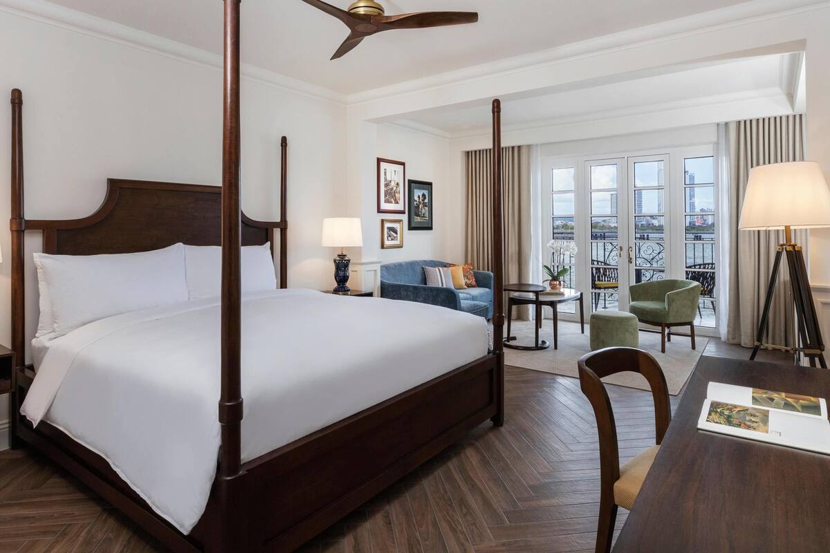 A hotel room with a four-poster bed, a ceiling fan, a balcony with a view, and a dining area with a table and chairs.