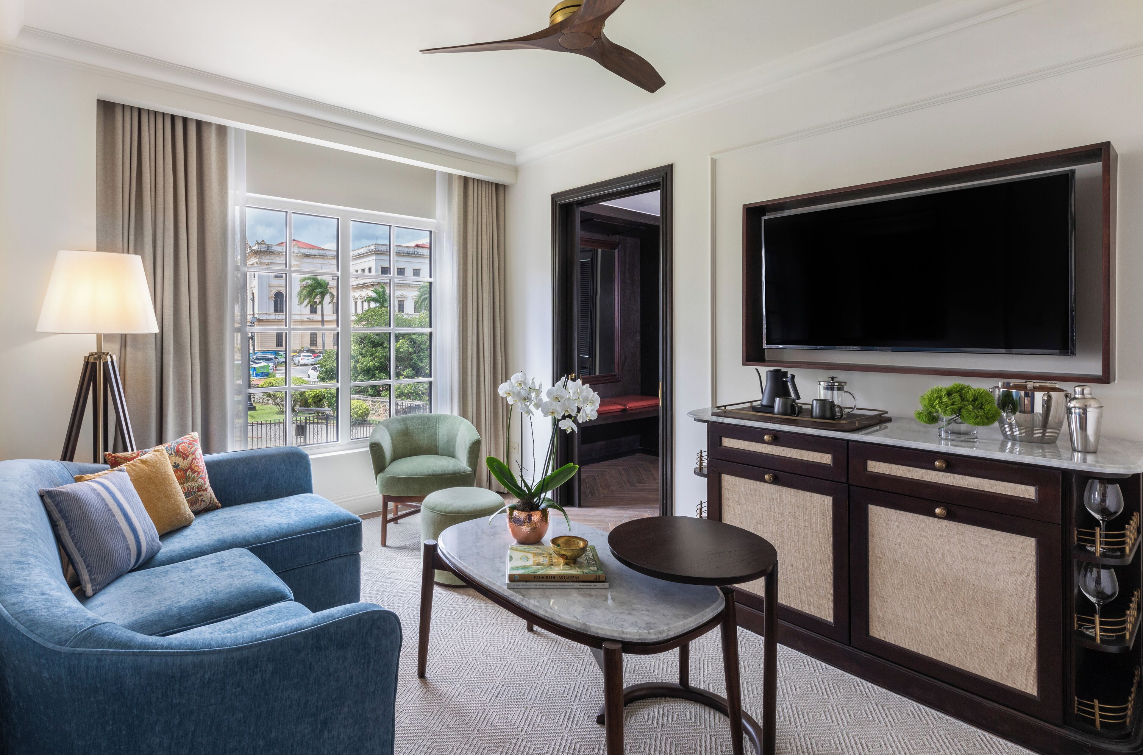 A hotel room with a blue sofa, a small table with a book and a plant, a TV, and a view of buildings outside.