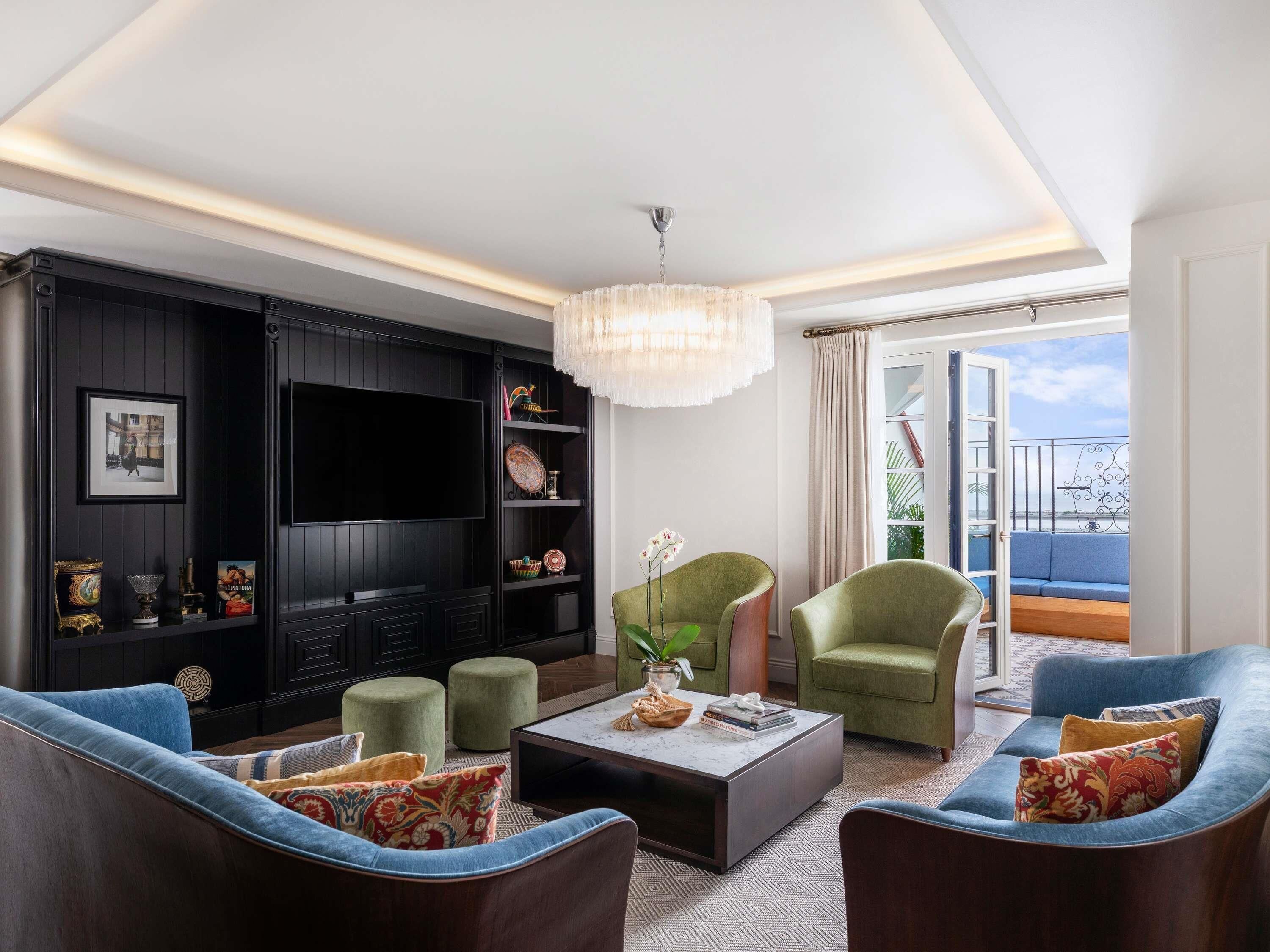 A modern living room with a flat-screen TV, green armchairs, a coffee table, and a balcony with a view.