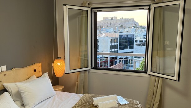A hotel room with a bed, a nightstand, a lamp, and a window with an open view of a cityscape and a historical landmark. - Park Minimal (Marousi)