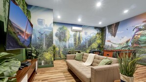 A modern living room with a large flat-screen TV, a beige sofa, wooden flooring, and a mural of a forest scene.