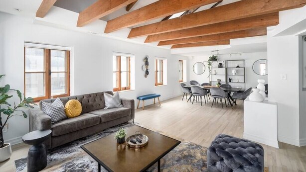 A spacious living area with a wooden ceiling, a grey sofa, a coffee table, and a dining area in the background. - Sublime 3BR 2BA Apt | Old Mtl | St Paul ST | (Montréal)