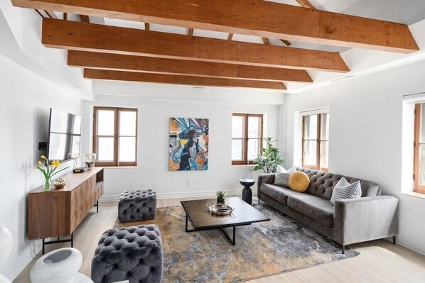 Un salon spacieux avec un plafond haut, des poutres apparentes, un canapé gris, une table basse et un tableau accroché au mur.