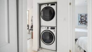 A laundry room with two stacked washing machines, a bed with white bedding, and a painting on the wall. - Chic & Modern 3B 1B Apt | Old Mtl | St Paul St | (Montréal)