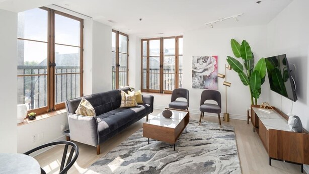 A spacious living room with large windows, a sofa, chairs, a coffee table, and a TV mounted on the wall. - Chic & Modern 3B 1B Apt | Old Mtl | St Paul St | (Montréal)