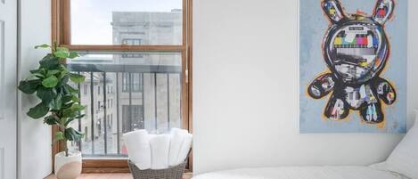 A bedroom with a large window, a potted plant, a basket of towels, and a colorful abstract painting on the wall.
