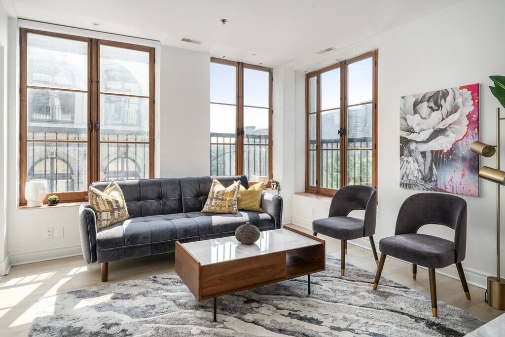 Ein modernes Wohnzimmer mit einem dunkelblauen Sofa, einem Holztisch und zwei grauen Sessel. Große Fenster bieten Blick auf die Stadt.