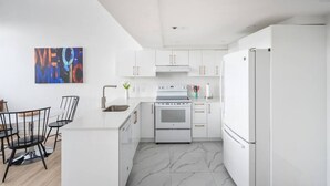 Une cuisine moderne avec des armoires blanches, un four en acier inoxydable et un réfrigérateur blanc. On y trouve un coin repas avec des chaises noires et une table blanche.