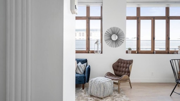 Un salon avec une grande fenêtre, un canapé bleu, un fauteuil marron et un miroir rond accroché au mur.