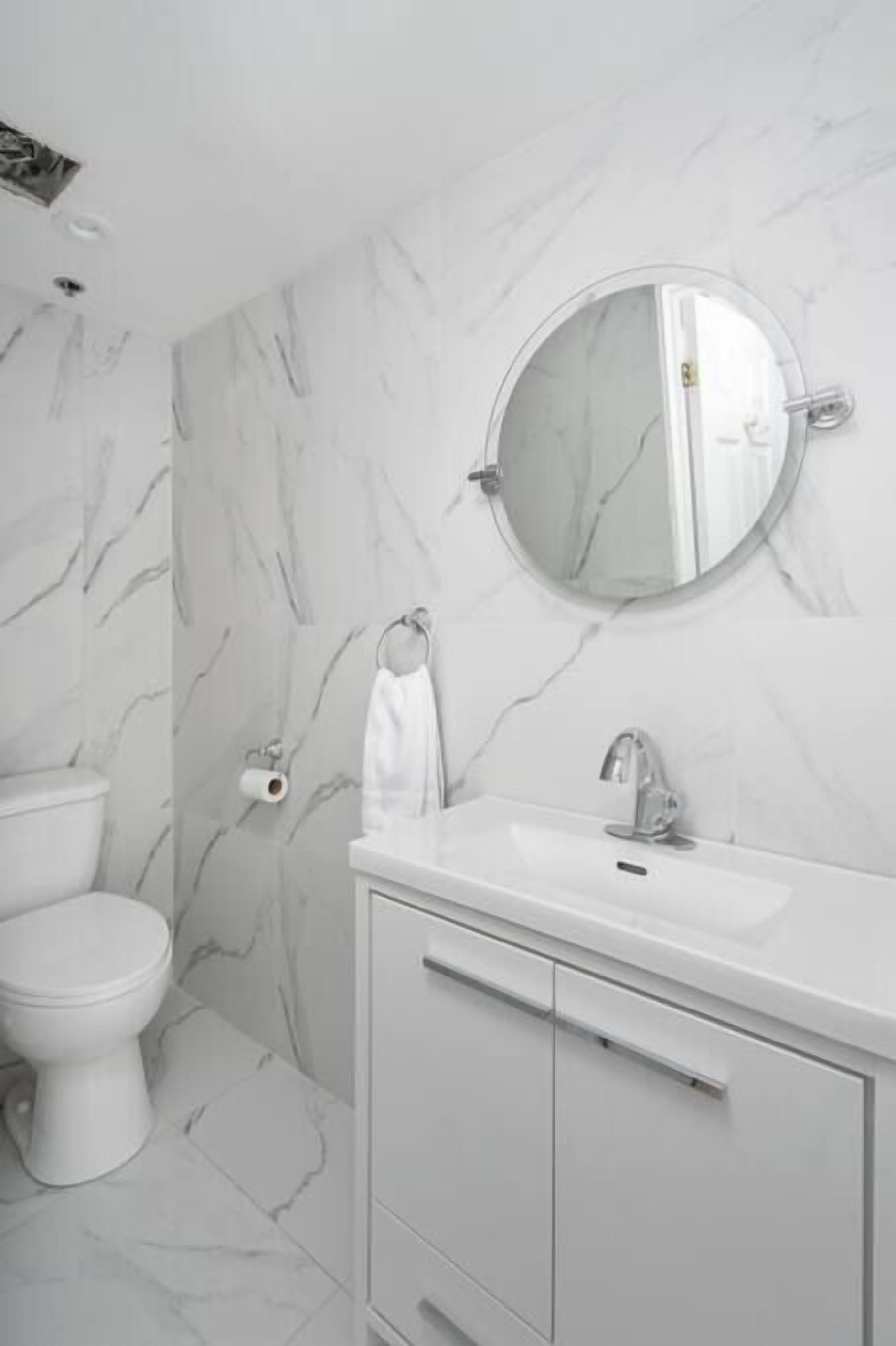 Un baño con inodoro blanco, lavamanos con espejo arriba y paredes con diseño de mármol.