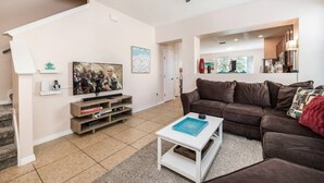 Een woonkamer met een bruine leren bank, een witte salontafel, een televisie op een houten dressoir, en een trap die naar een andere kamer leidt.