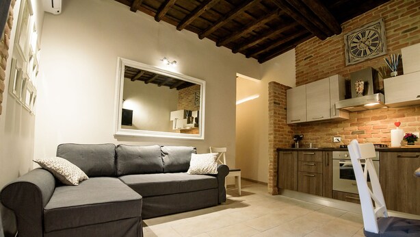 A living room with a grey sofa, a white chair, and a kitchen area with wooden cabinets and a brick wall. - NEW NAVONA ELEGANT APARTMENT (Roma)