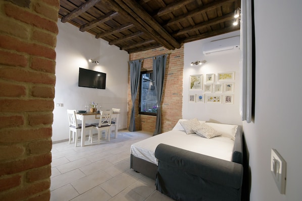 Quarto com cama, escrivaninha e cadeira, televisão presa à parede e janela com cortinas.
