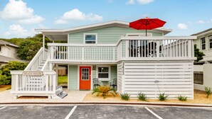 A two-story beach house with a red door, a balcony with a red umbrella, and a small patio area with plants. - Tropical Beach Bungalow Up | 3 bedrooms | Across Street from Beach | Deck (Fernandina Beach)