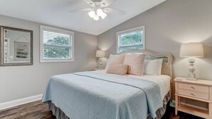 A bedroom with a bed, two bedside tables, a ceiling fan, and two windows with blinds. - Tropical Beach Bungalow Up | 3 bedrooms | Across Street from Beach | Deck (Fernandina Beach)