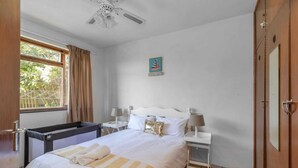 A bedroom with a bed, two bedside tables, a ceiling fan, a window with curtains, and a door. - Sunny Tides Holiday Inn (Kingsburgh)