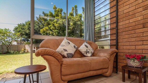 A tan leather sofa with leaf-patterned cushions, a small round table, and a flower vase on a tiled patio with a brick wall and large windows. - Sunny Tides Holiday Inn (Kingsburgh)