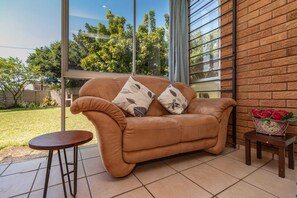 A tan leather sofa with leaf-patterned cushions, a small round table, and a flower vase on a tiled patio with a brick wall and large windows.