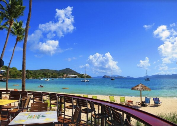 Un restaurante frente a la playa con terraza, palmeras y vistas al mar con barcos.