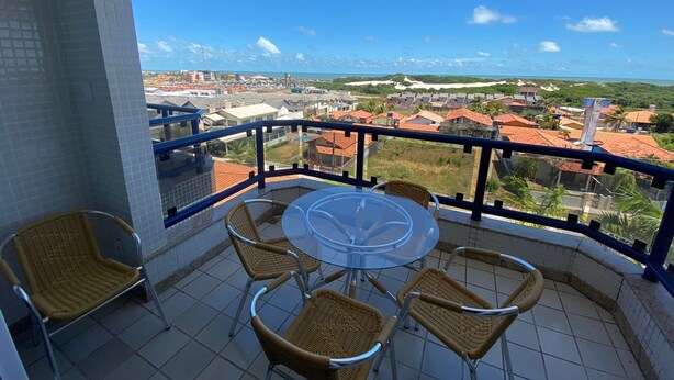 A balcony with a glass table and wicker chairs, overlooking a coastal town with red-roofed houses and a clear blue sky. - Calypso solar building A gated community in Atalaia close to the beach. (salinopolis)