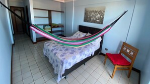 A bedroom with a bed, a hammock, a chair, a shelf with folded clothes, and a cityscape painting on the wall. - Calypso solar building A gated community in Atalaia close to the beach. (salinopolis)