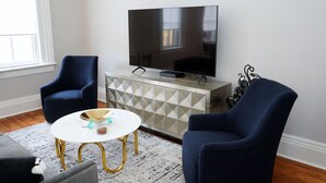 Una sala de estar con dos sillones azules, una mesa de centro blanca con patas doradas, un televisor de pantalla plana sobre un mueble con estampado geométrico, y una ventana con persianas.