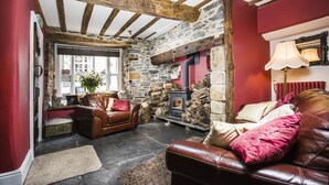 A cozy living room with a stone fireplace, wooden beams, a leather sofa, and a red wall. - Cornerstones - 16th Century House overlooking River in Llangollen (LLANGOLLEN)