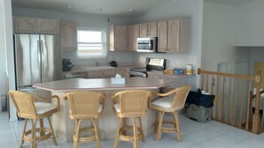 A kitchen with a central island, four wicker chairs, stainless steel appliances, and a wooden staircase.