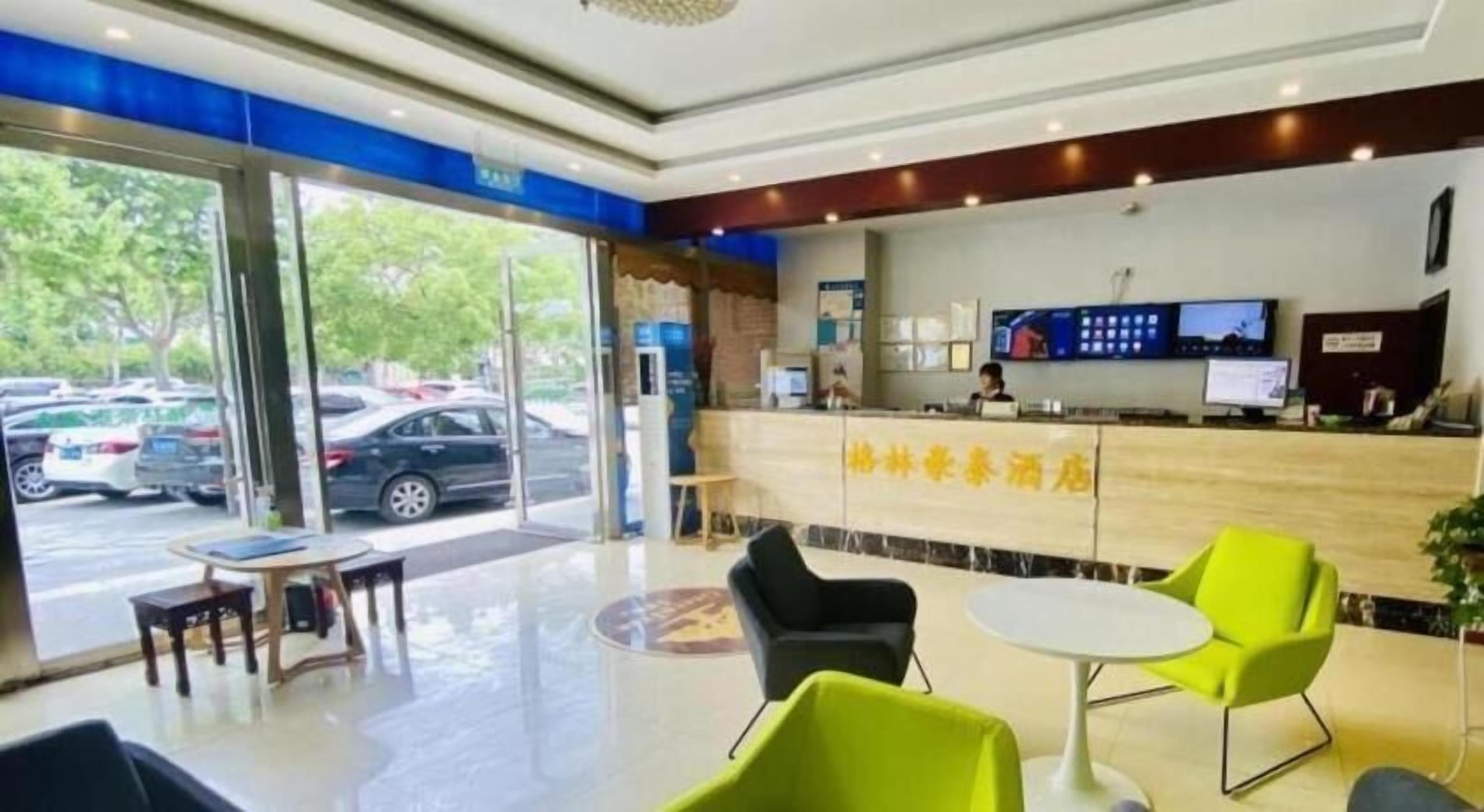 Un hall de hotel con mostrador de recepción, zona de estar y grandes ventanales con vistas a coches estacionados.