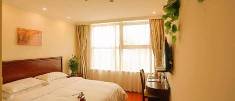 A hotel room with a bed, a desk, a chair, a window with curtains, a plant, and an air conditioning unit.