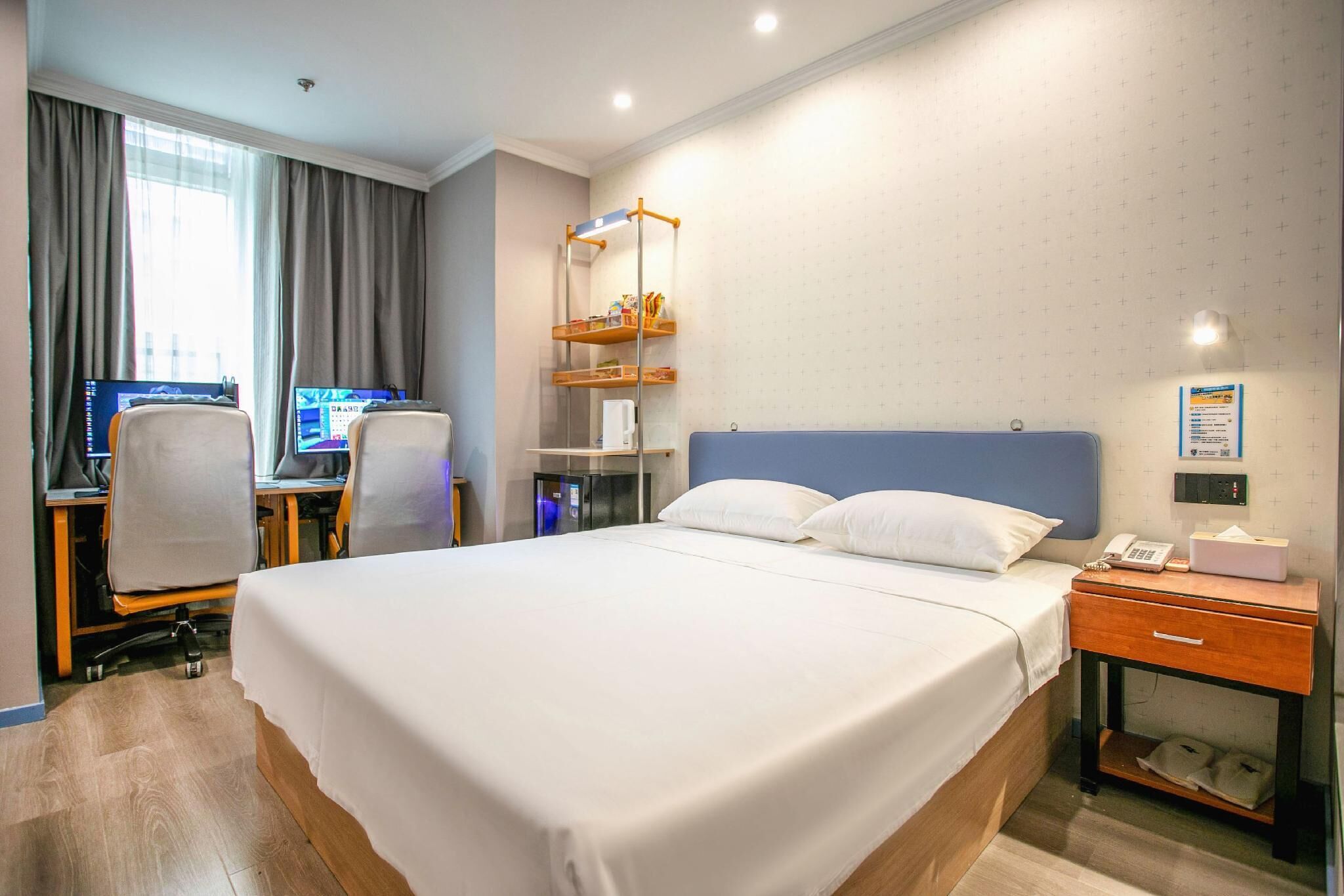 Quarto de hotel com uma cama grande, uma mesa com dois monitores, uma cadeira, prateleira com artigos de higiene e mesa de cabeceira com telefone e lenços.
